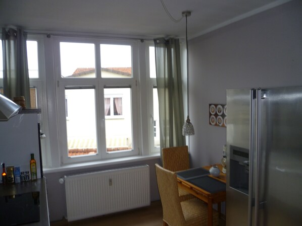 Een kleine keuken met een eettafel, stoelen, een koelkast en een raam voorzien van gordijnen.