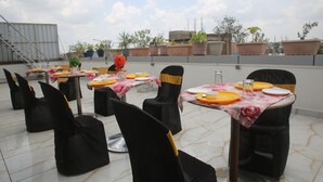 A rooftop dining area with tables set for four, black chairs with yellow sashes, and potted plants in the background.