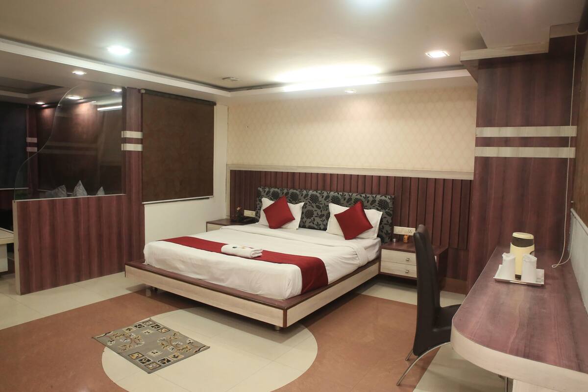 A modern hotel room with a large bed, a bedside table, a mirror, and a small desk with a chair.