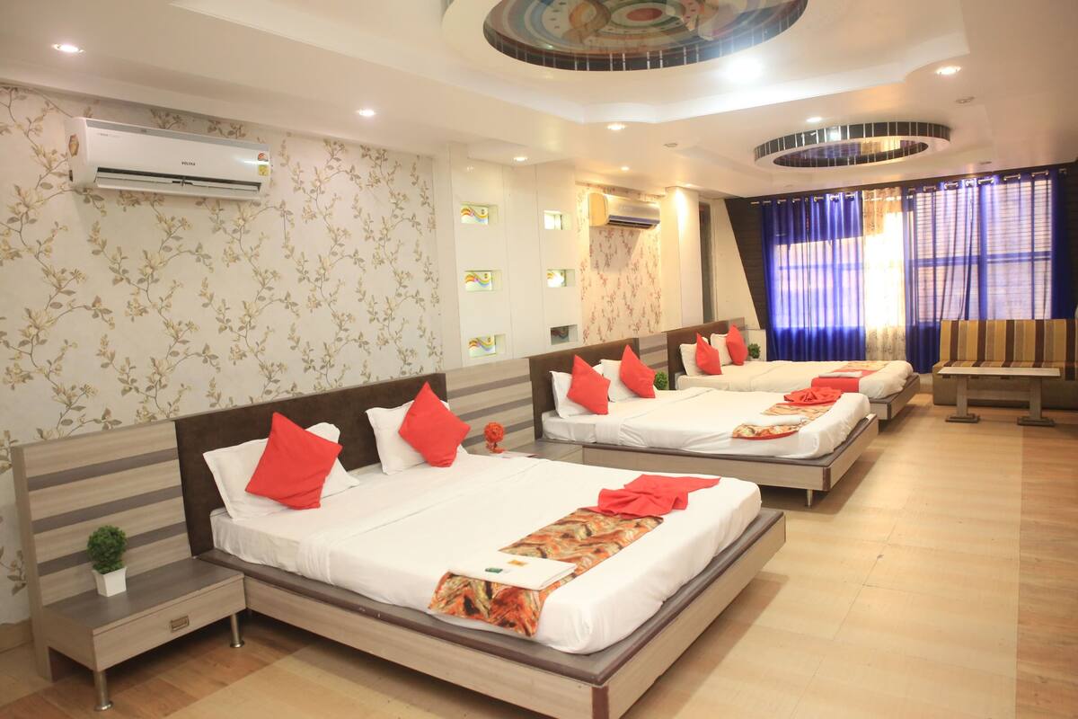 A hotel room with four beds, each with white bedding and red pillows, a small bedside table with a plant, and a wall-mounted air conditioner.