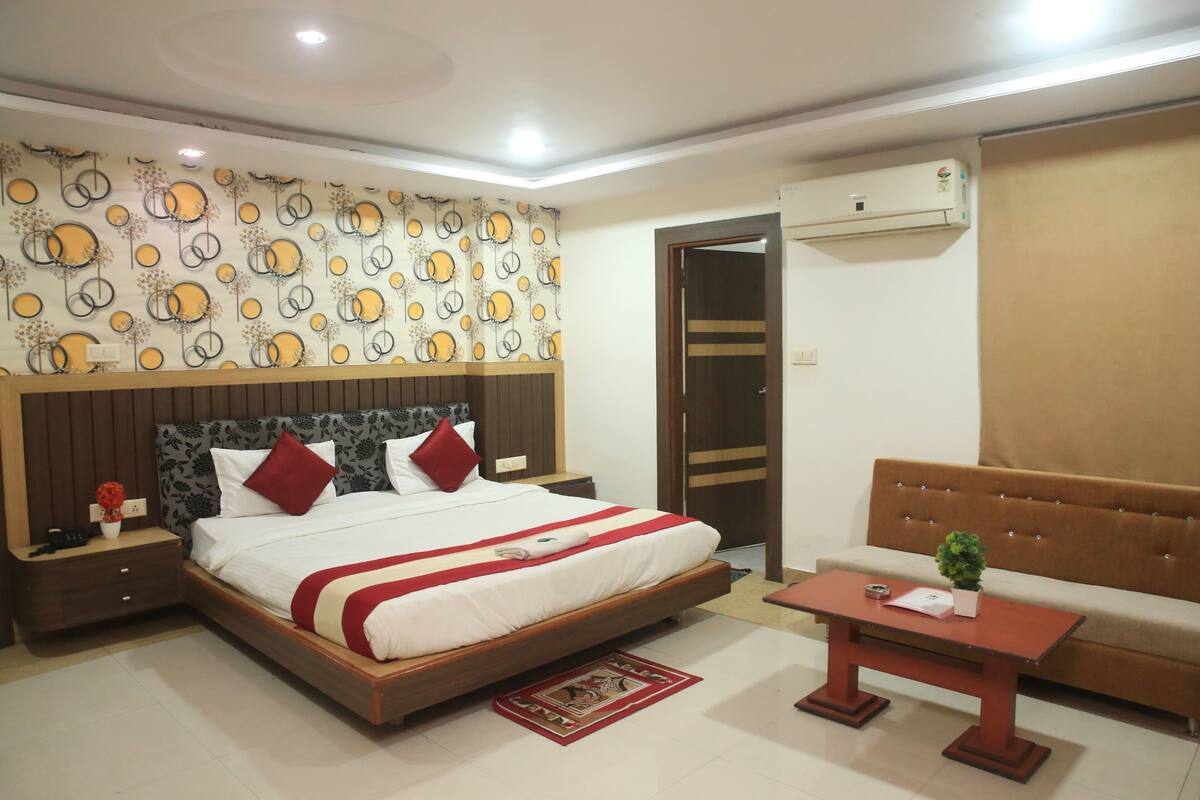 A hotel room with a bed, a nightstand, a table, a sofa, an air conditioning unit, and a patterned wall.
