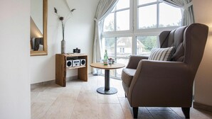 Un salon moderne avec une grande fenêtre, une table ronde, un fauteuil confortable, une étagère en bois avec une radio et un miroir.