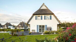 Une maison blanche avec un toit à forte pente et des volets bleus, entourée d’un jardin bien entretenu et d’un étang.