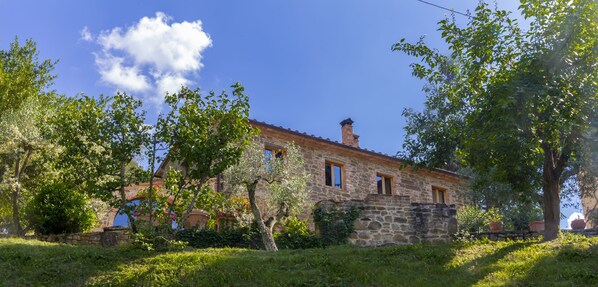 A stone building with a red-tiled roof, surrounded by lush greenery and trees.