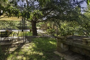 An outdoor patio area with a table and chairs, a grill, and a large tree providing shade.