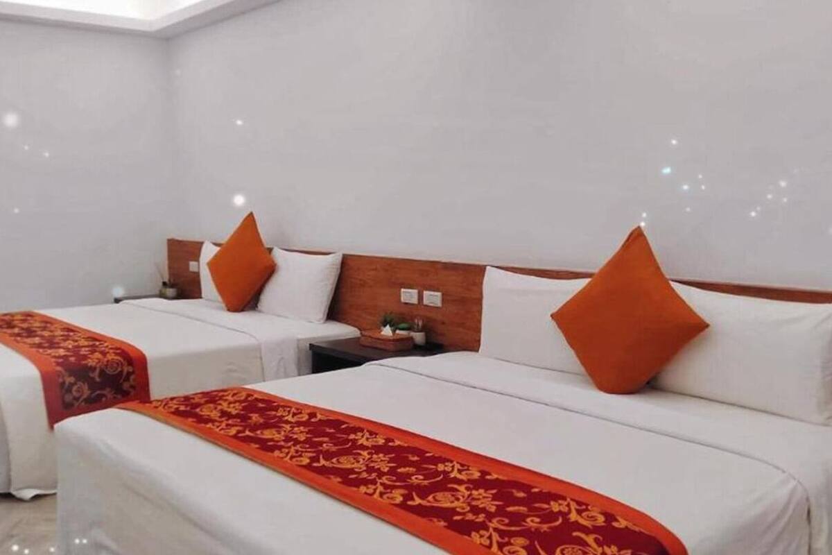 Habitación de hotel con dos camas, cada una con sábanas blancas y almohadas naranjas. Tiene un cabecero de madera y una mesita de noche con una lámpara.