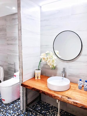 Une salle de bain avec un meuble-lavabo en bois, un miroir rond, un lavabo blanc, un petit vase avec des fleurs et un sol carrelé.