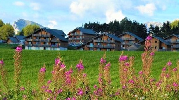 Eine Reihe von holzgebauten Chalets mit Balkonen vor einer Bergkulisse und blauem Himmel.