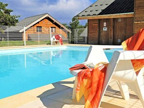 Une piscine entourée de chaises longues blanches, un chalet en bois et un espace délimité par une clôture.