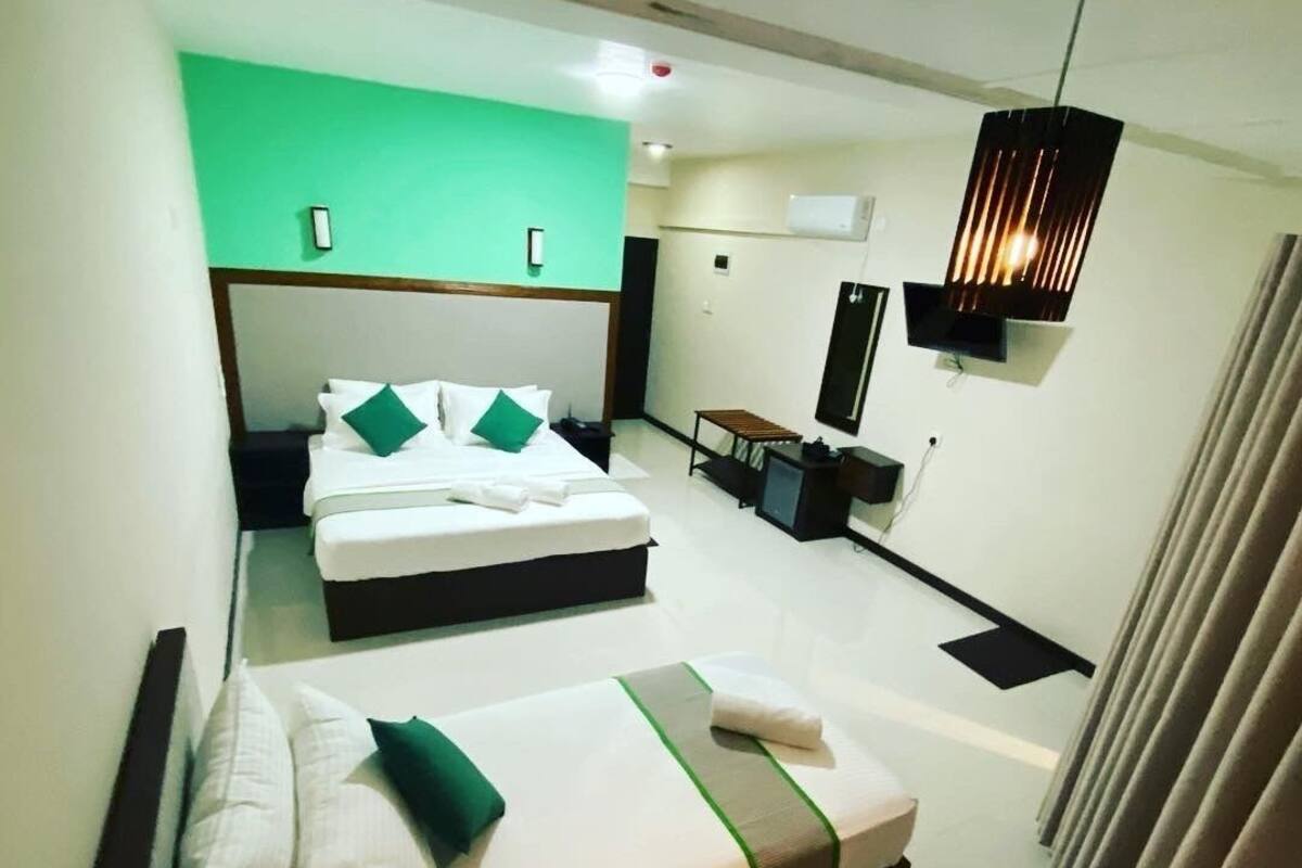 A hotel room with two beds, a green accent wall, a TV, a desk, and a hanging light fixture.