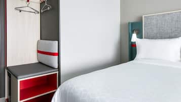 Una habitación de hotel con cama, un banco rojo y gris, un armario y una lámpara de pared.