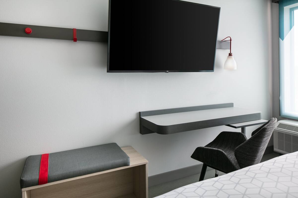 Ein modernes Hotelzimmer mit einem Wandfernseher, einem Schreibtisch, einem Stuhl, einer Bank mit rotem Akzent und einem Fenster mit Blick.