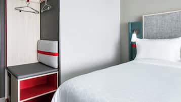 Quarto de hotel com cama, área de estar com um banco, guarda-roupa e luminária fixada na parede.