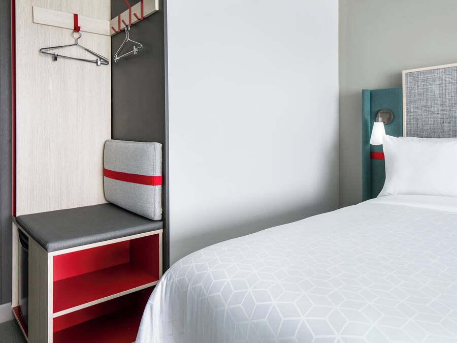 Quarto de hotel com cama, área de estar com um banco, guarda-roupa e luminária fixada na parede.