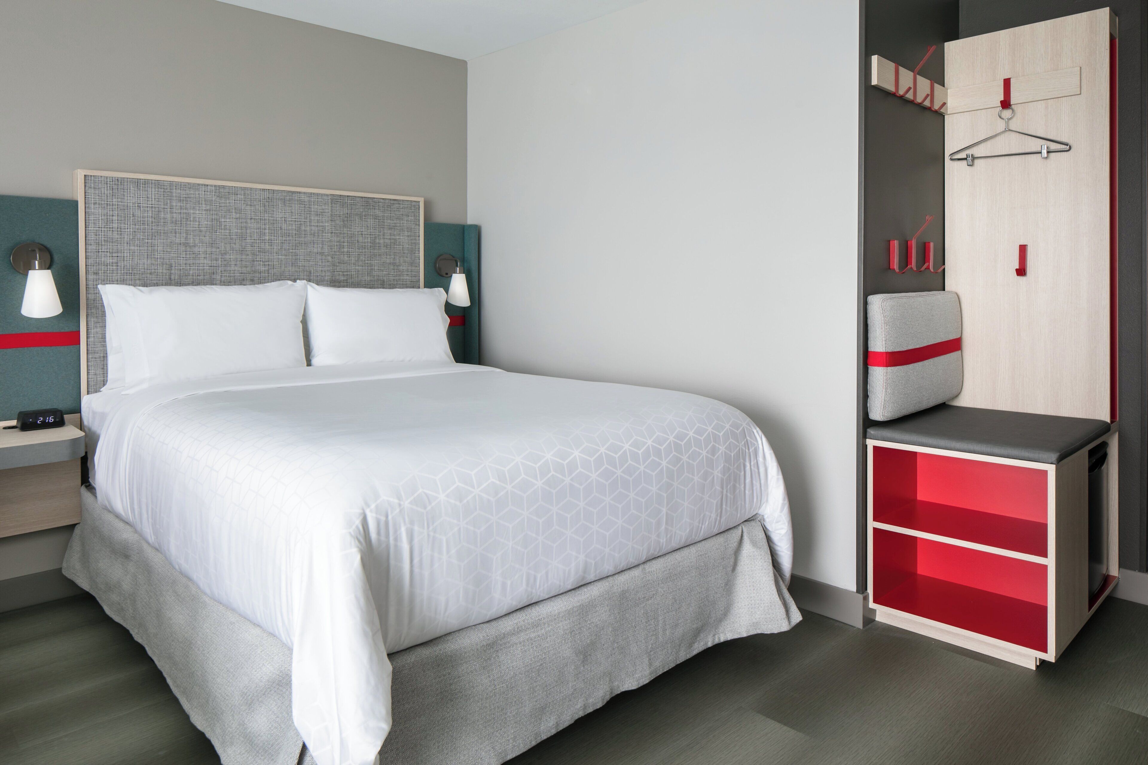 A modern hotel room with a large bed, a wardrobe with red accents, and a bedside table with a lamp.