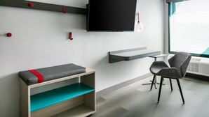 A modern room with a mounted TV, a bench with a red accent, a chair, and a window with a view. - avid hotel Roseville - Minneapolis North by IHG (Roseville)