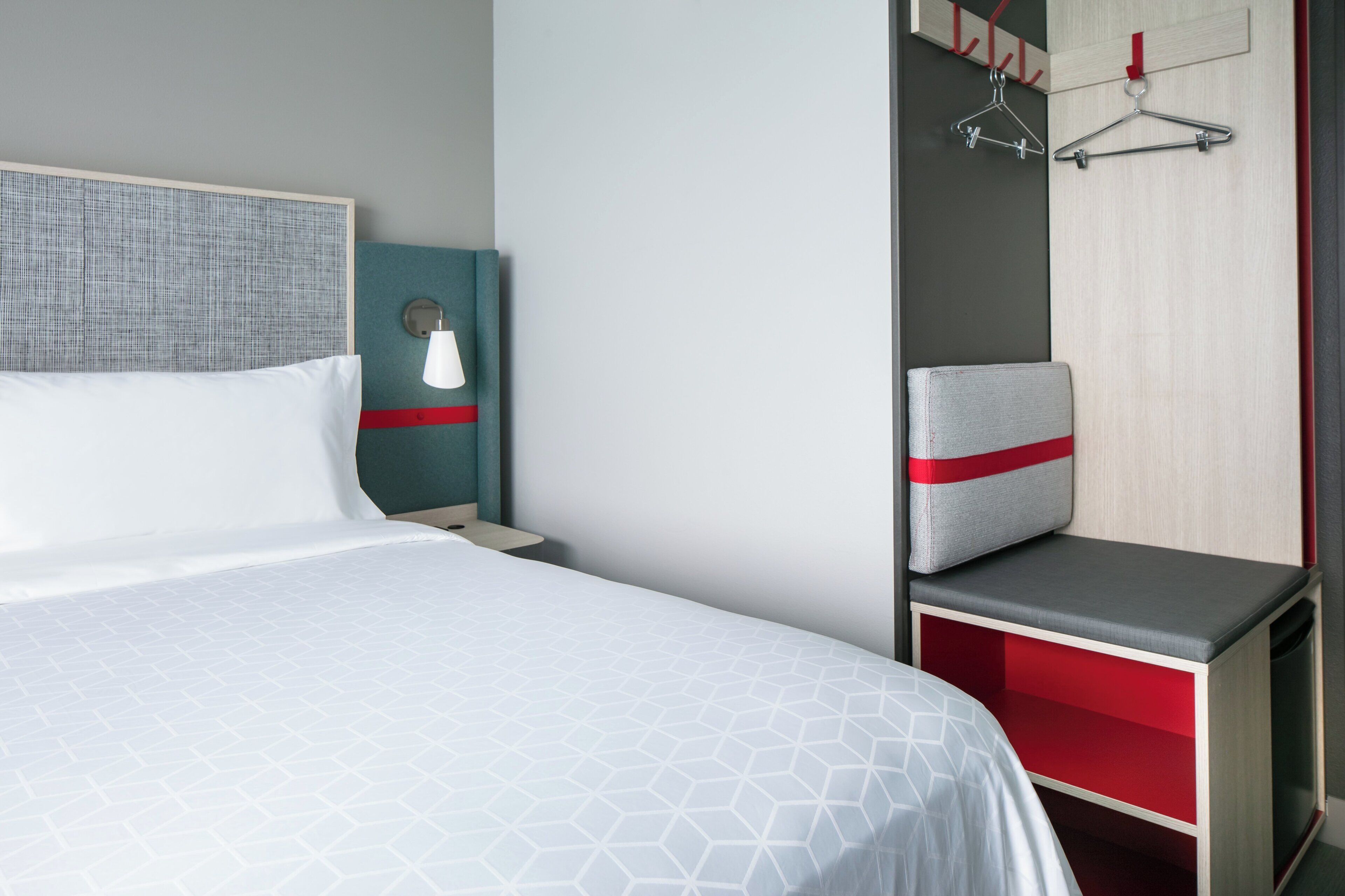 Habitación de hotel con una cama grande, una mesita de noche, una lámpara de pared, una zona de estar con un banco y un perchero.