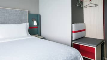 Habitación de hotel con una cama grande, una mesita de noche, una lámpara de pared, una zona de estar con un banco y un perchero.