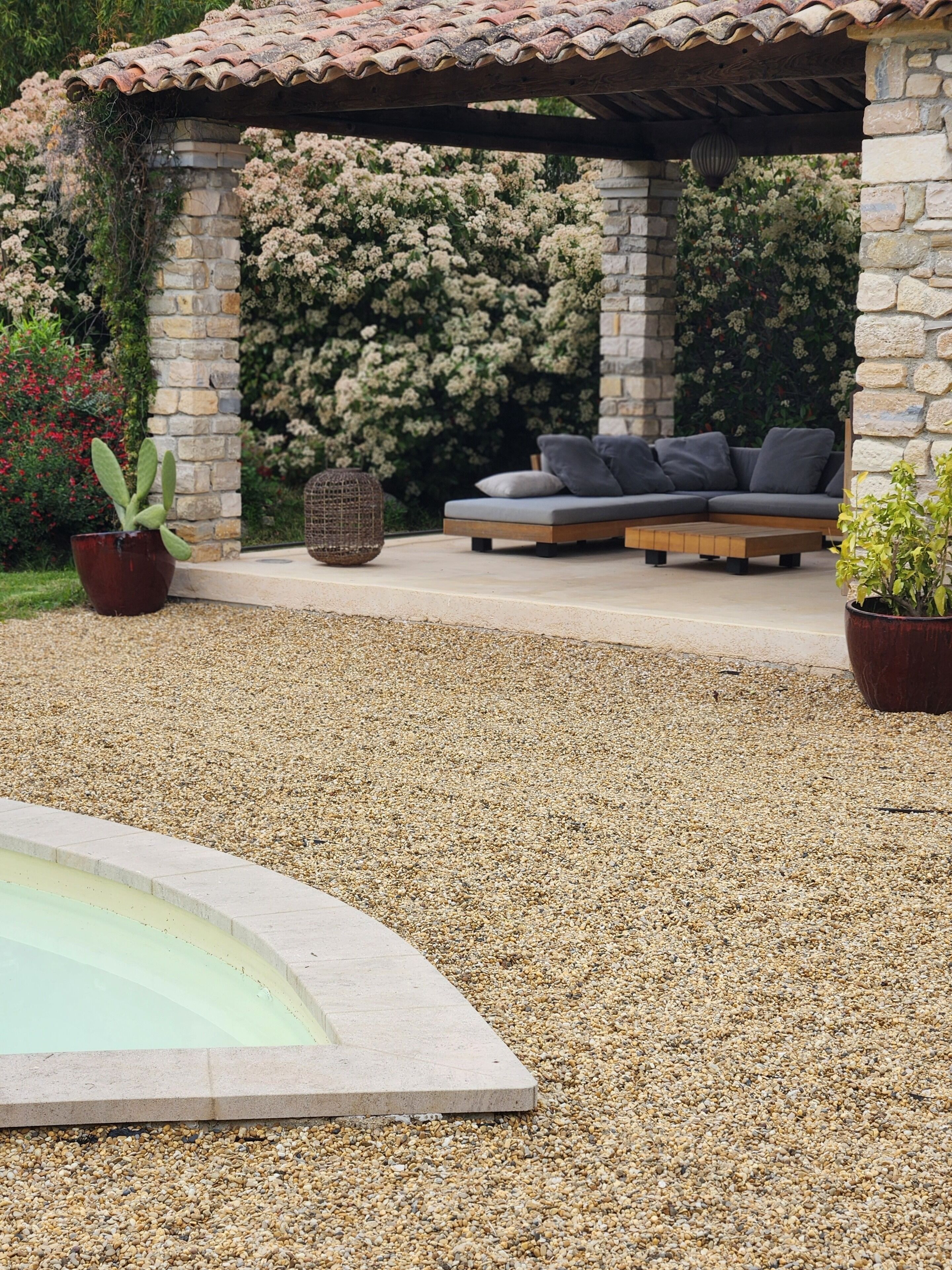 A covered outdoor seating area with a stone wall and a tiled roof, surrounded by gravel and a pool.