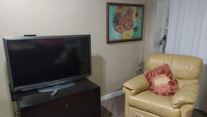 Un salon avec un téléviseur à écran plat posé sur un meuble en bois foncé, un fauteuil en cuir beige agrémenté d’un coussin à motifs et un tableau représentant des tournesols accroché au mur.
