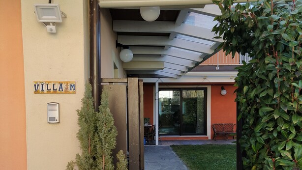 Une entrée d’immeuble avec une enseigne indiquant « VILLAM », une plante en pot et un chemin menant à un espace aménagé avec des sièges.