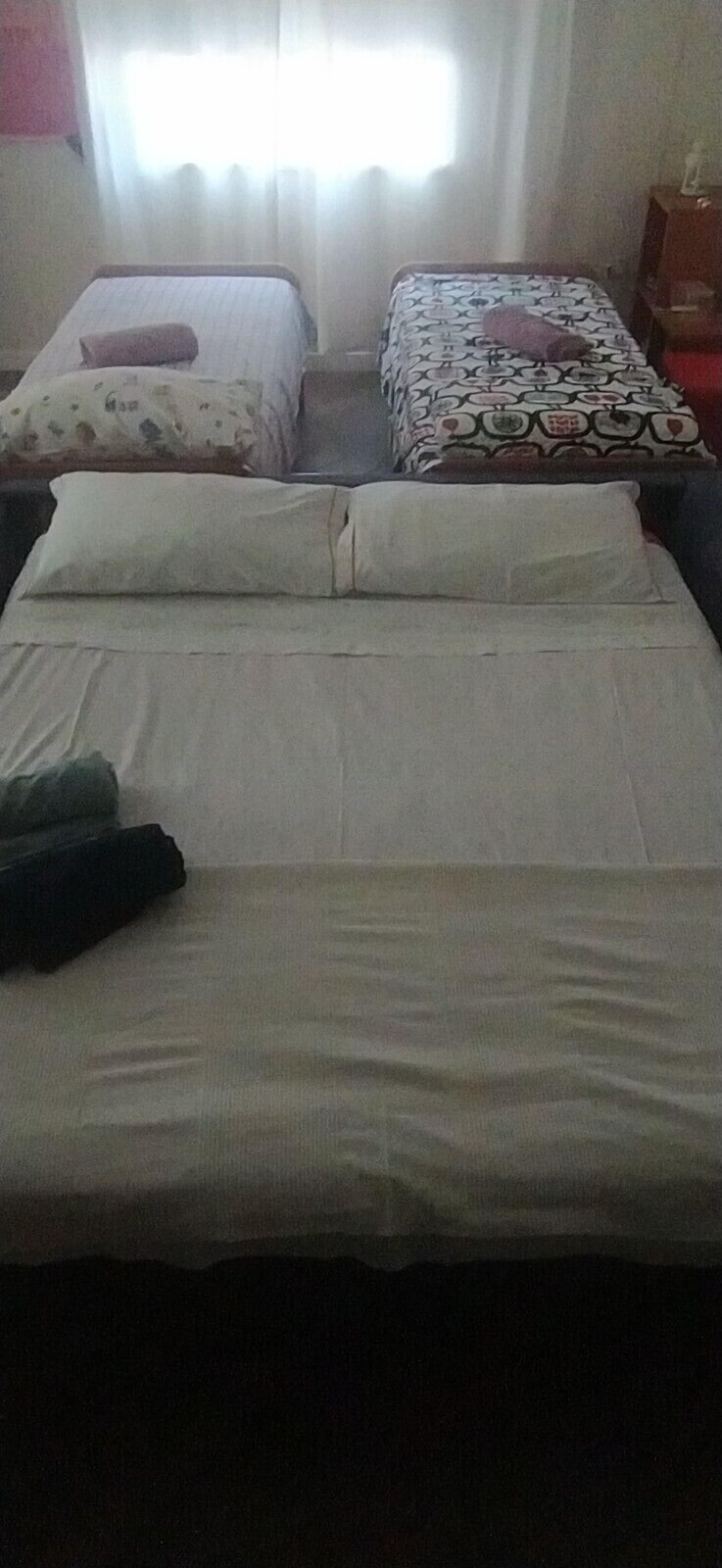 A hotel room with two beds, one with a patterned bedspread and the other with a plain one. There is a window with curtains, a nightstand, and a lamp.
