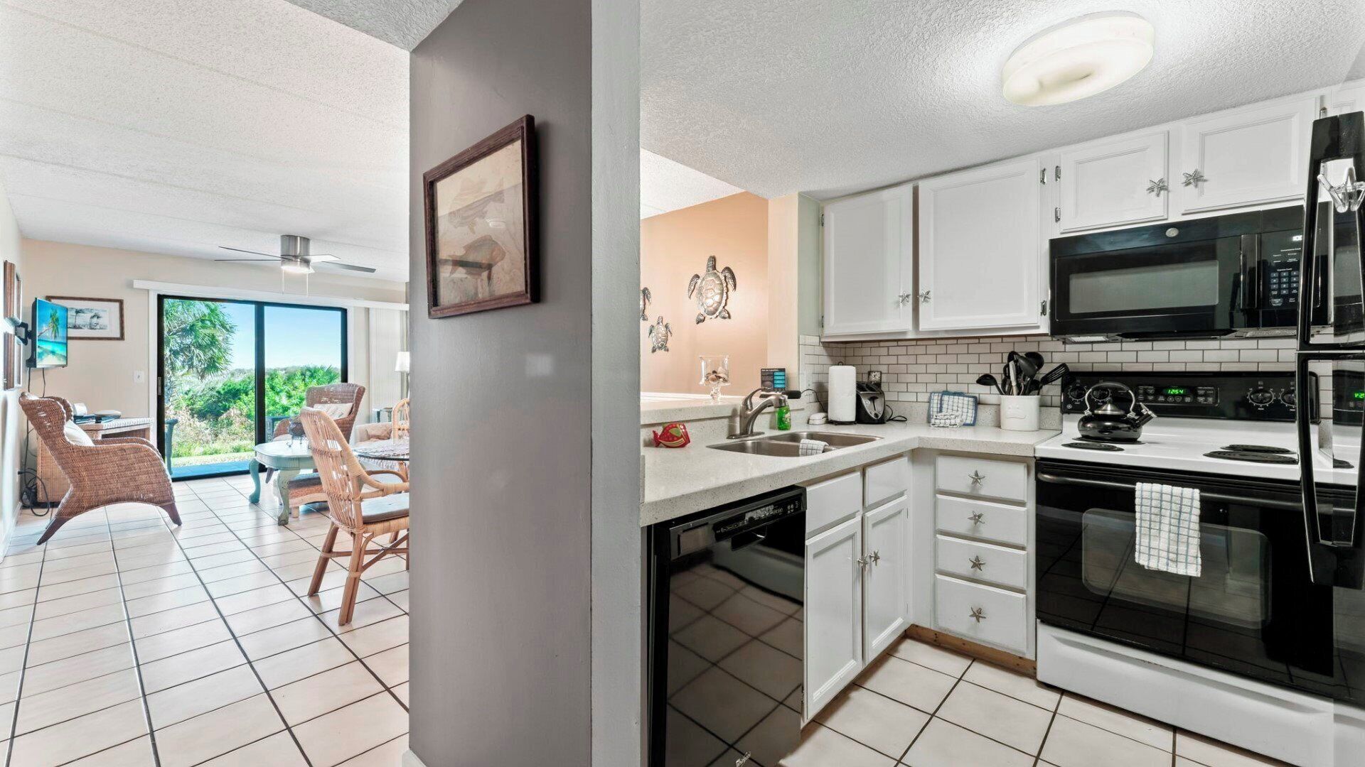 A modern kitchen with white cabinets, a black microwave, and a stove. There is a dining area with wicker chairs and a view of greenery outside.