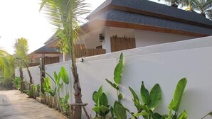 A pathway lined with banana plants and a white wall, leading to a building with a dark roof and wooden fence.