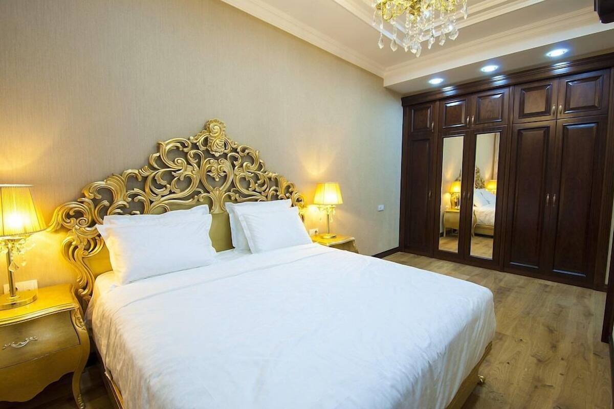 A hotel room with a large bed, ornate headboard, two bedside lamps, a chandelier, and a wardrobe with mirrors.