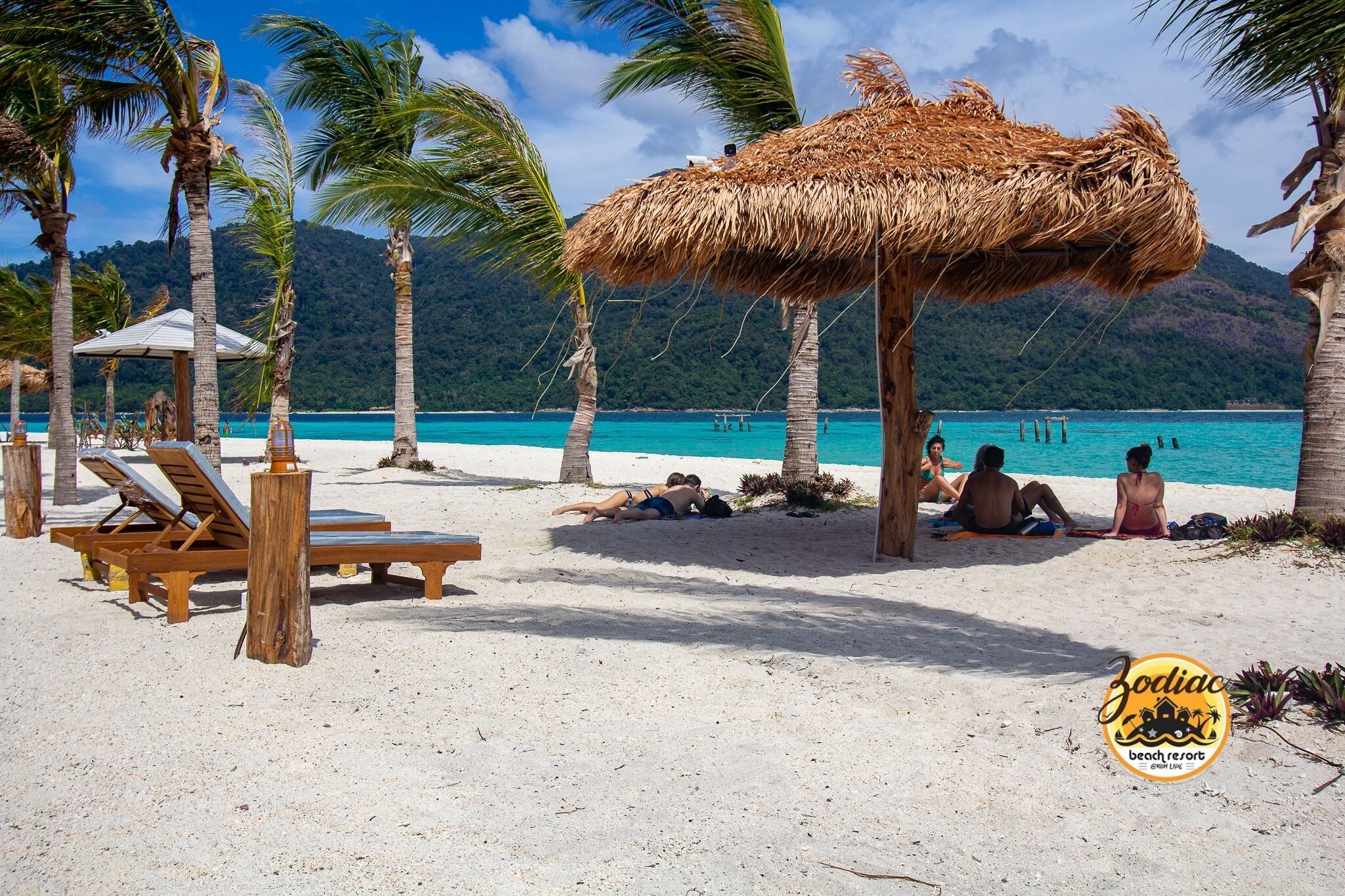 A beach with wooden lounge chairs, a thatched umbrella, and palm trees.