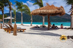 A beach with wooden lounge chairs, a thatched umbrella, and palm trees.