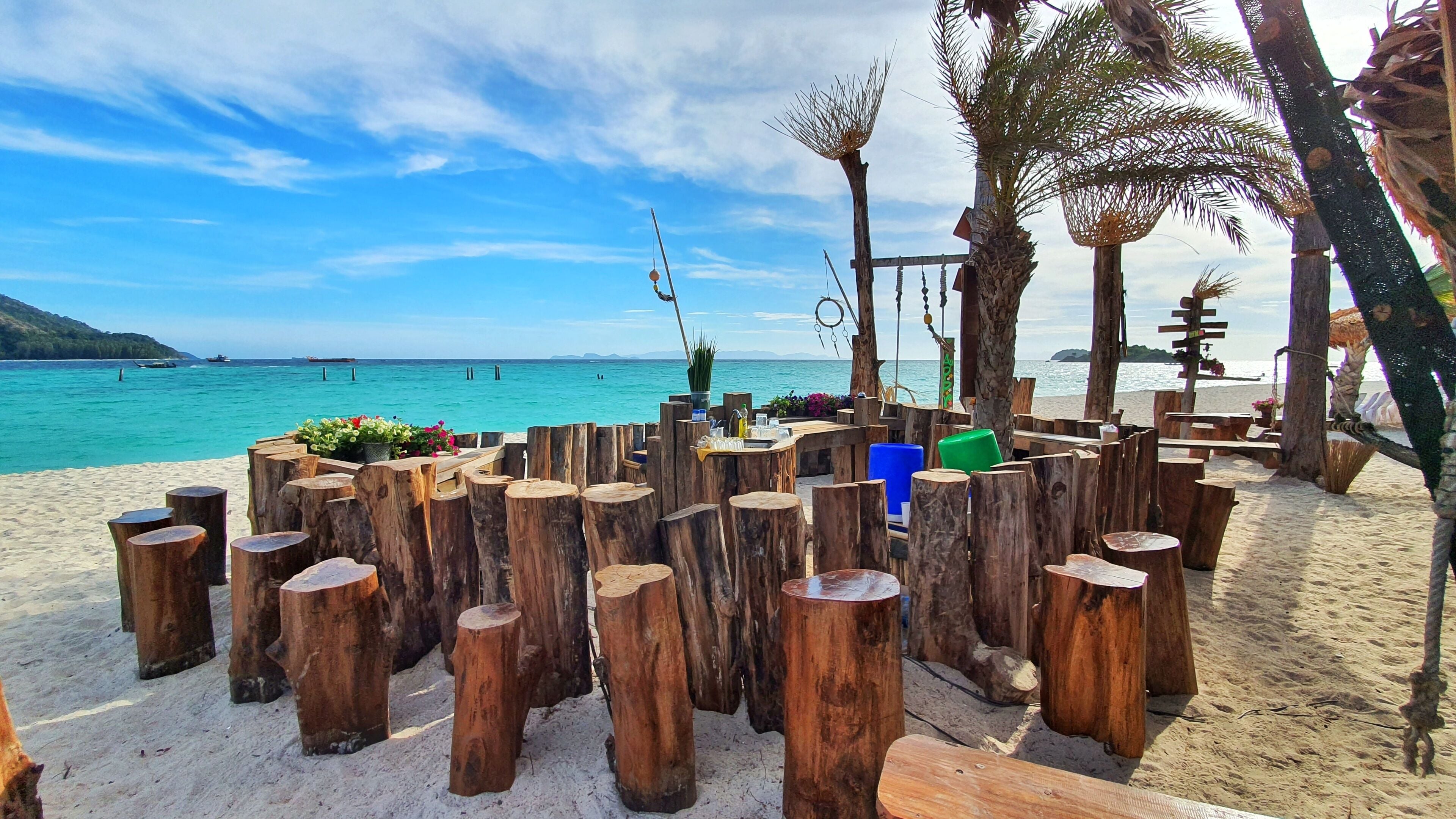 A beachside wooden structure with a bench and a table, surrounded by palm trees and a view of the sea.