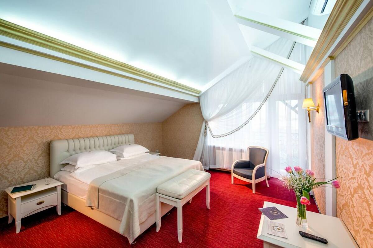 Ein Hotelzimmer mit einem großen Bett, einem Nachttisch, einem Sessel, einem Schreibtisch mit Lampe und einem an der Wand montierten Fernseher.