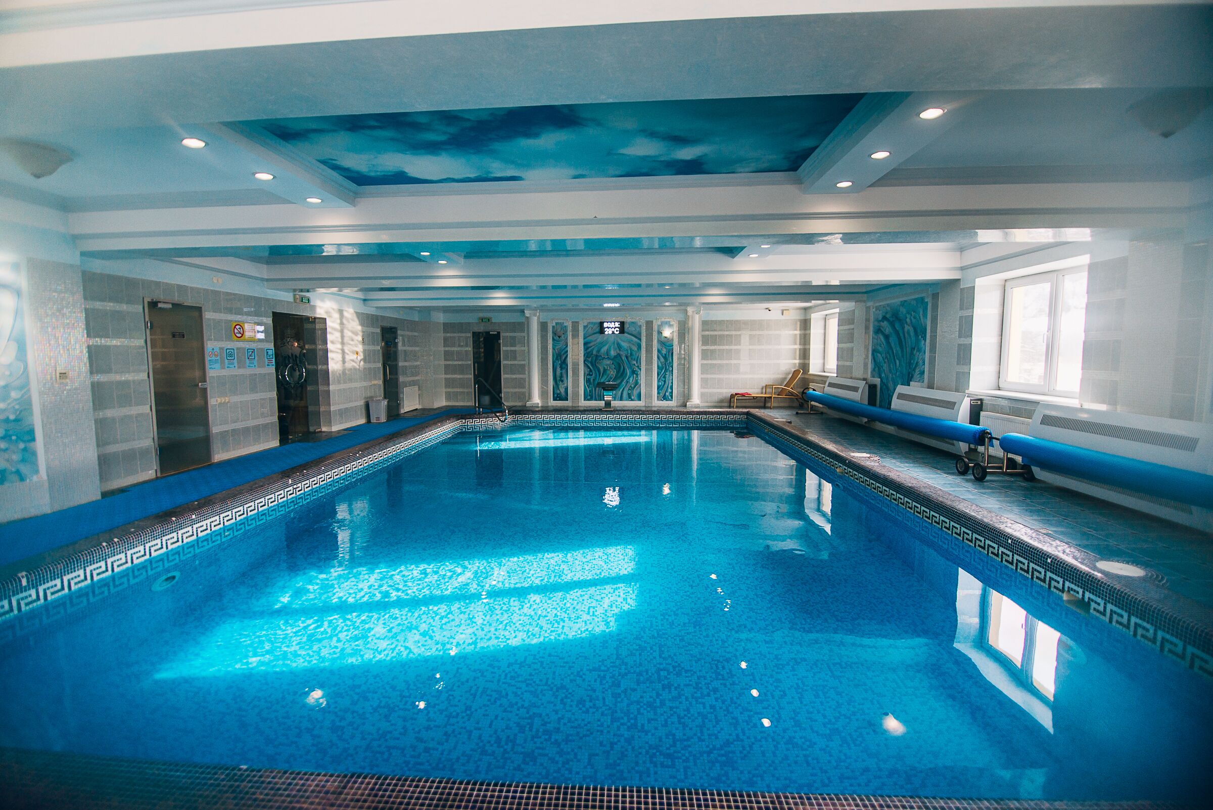 A large indoor swimming pool with a blue mosaic tile design, surrounded by walls with a geometric pattern and a ceiling with a cloud mural.