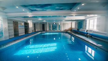 A large indoor swimming pool with a blue mosaic tile design, surrounded by walls with a geometric pattern and a ceiling with a cloud mural.