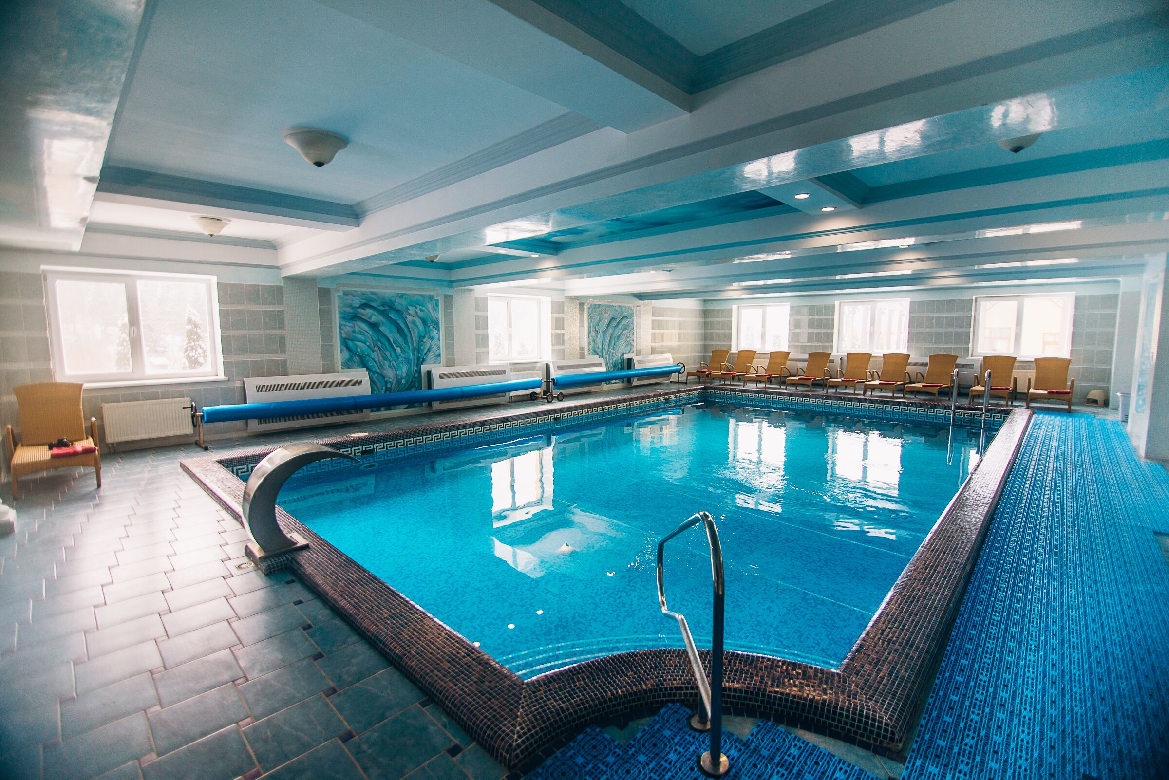 An indoor swimming pool with a blue tiled floor and walls, a diving board, and several chairs around the pool.