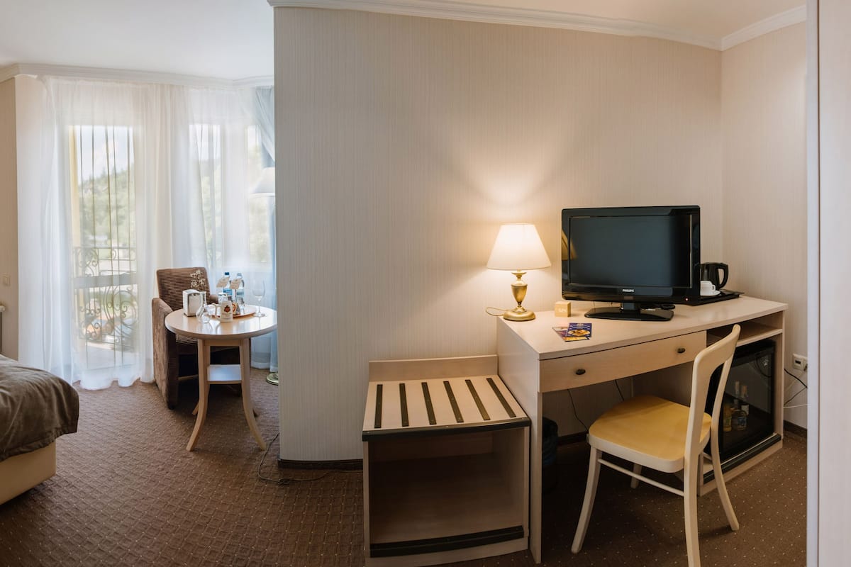 Ein Hotelzimmer mit Bett, Schreibtisch mit Computer, Stuhl, kleinem Tisch mit Flasche und Glas sowie einem Fenster mit Vorhängen.