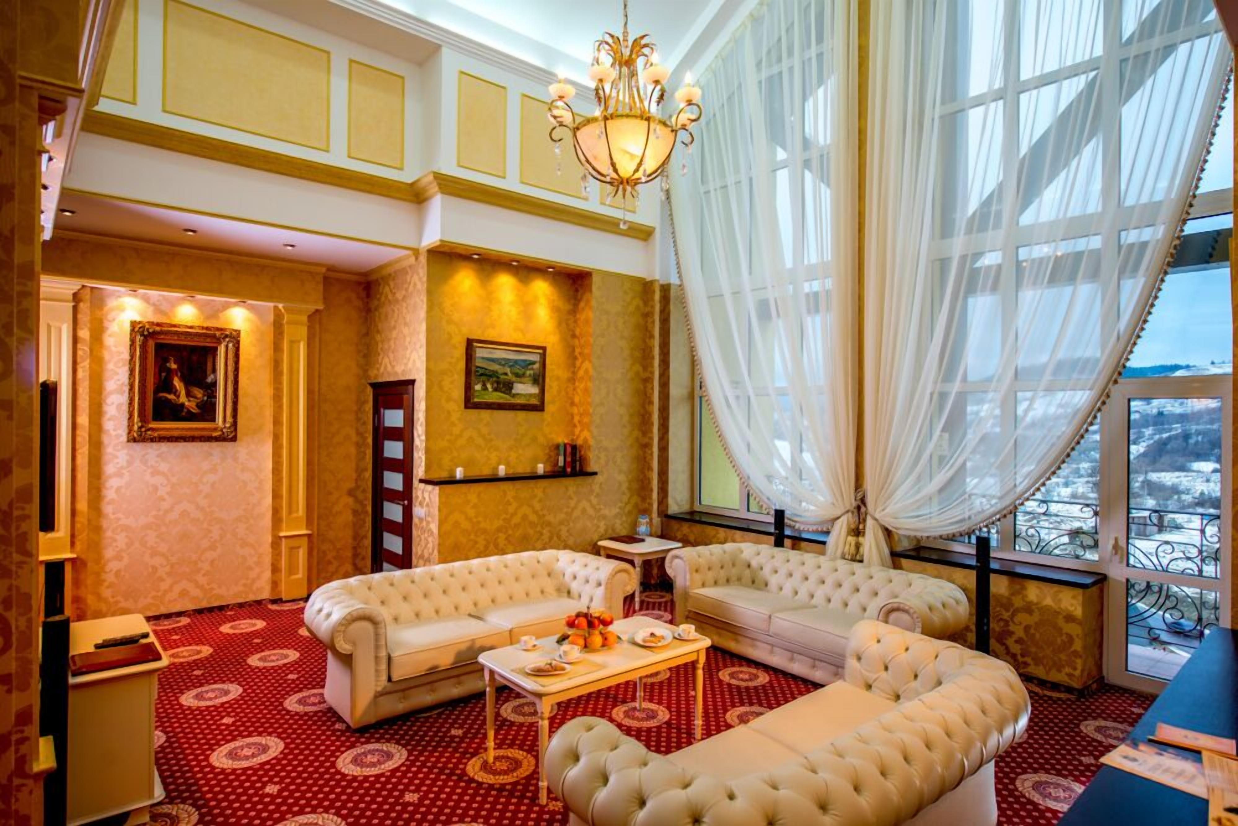 Ein luxuriöses Hotelzimmer mit einem roten Teppich mit Muster, zwei weißen, gepolsterten Sofas, einem Kronleuchter und einem großen Fenster mit durchsichtigen Vorhängen.