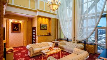 Ein luxuriöses Hotelzimmer mit einem roten Teppich mit Muster, zwei weißen, gepolsterten Sofas, einem Kronleuchter und einem großen Fenster mit durchsichtigen Vorhängen.