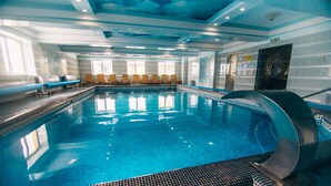 Una piscina interior con agua azul cristalina, paredes con azulejos y un techo con diseño azul y blanco. Hay sillas de descanso a lo largo de la piscina y un letrero que indica la zona de la piscina.