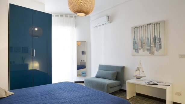 A bedroom with a large bed, a blue wardrobe, a small armchair, a bedside table with a book, and a wall-mounted air conditioner.