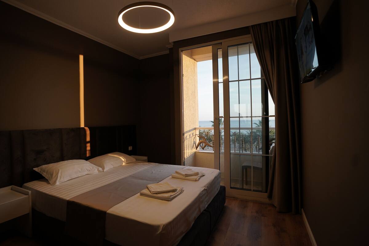 A modern hotel room with a large bed, bedside tables, a flat-screen TV mounted on the wall, and a balcony with a view.