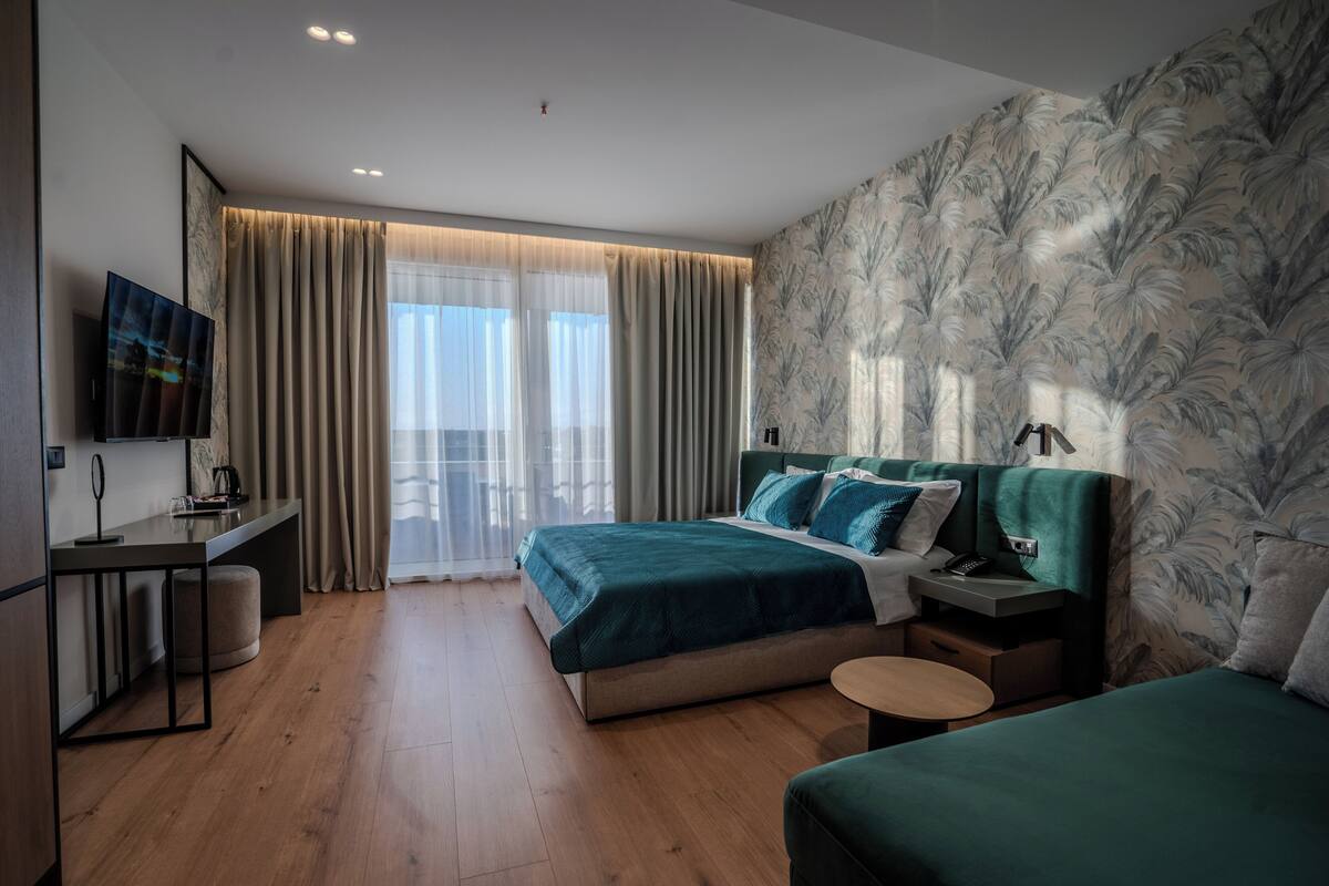 A modern hotel room with a large bed, a desk, a TV, and a balcony with curtains.