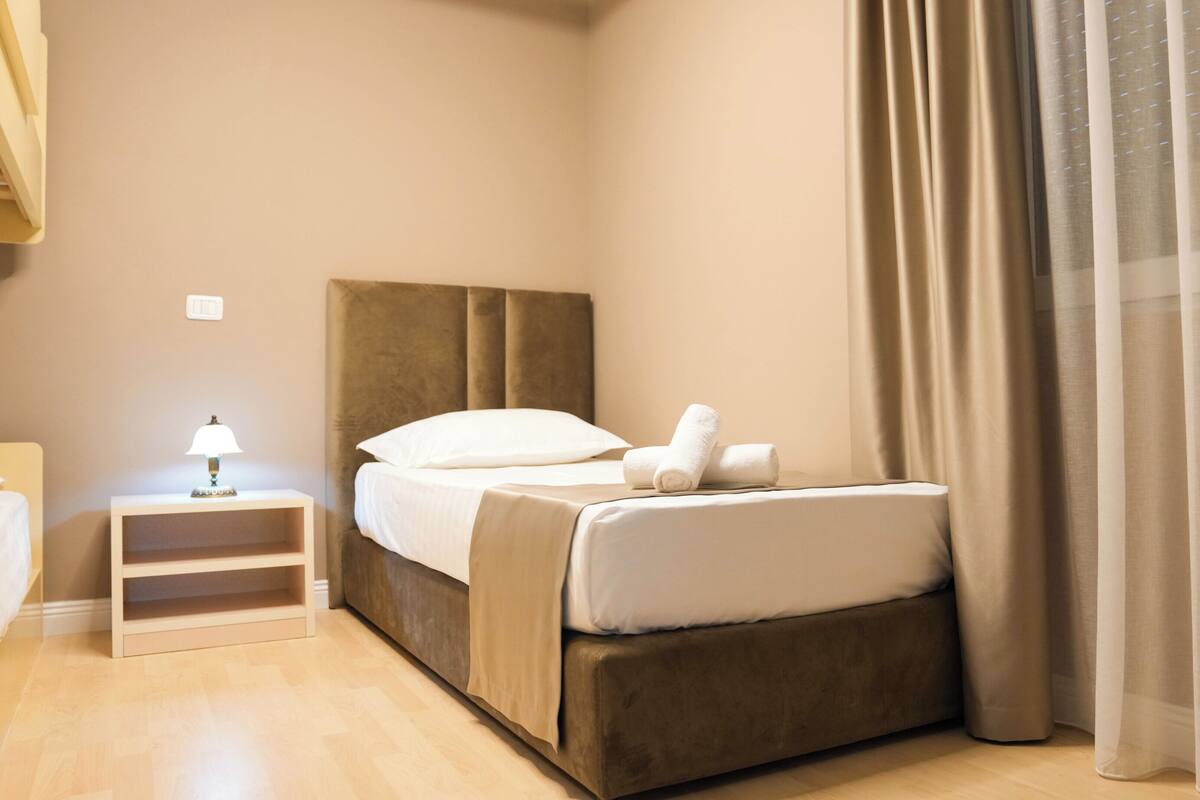 A neatly made bed with a headboard, a bedside table with a lamp, and a window with curtains.