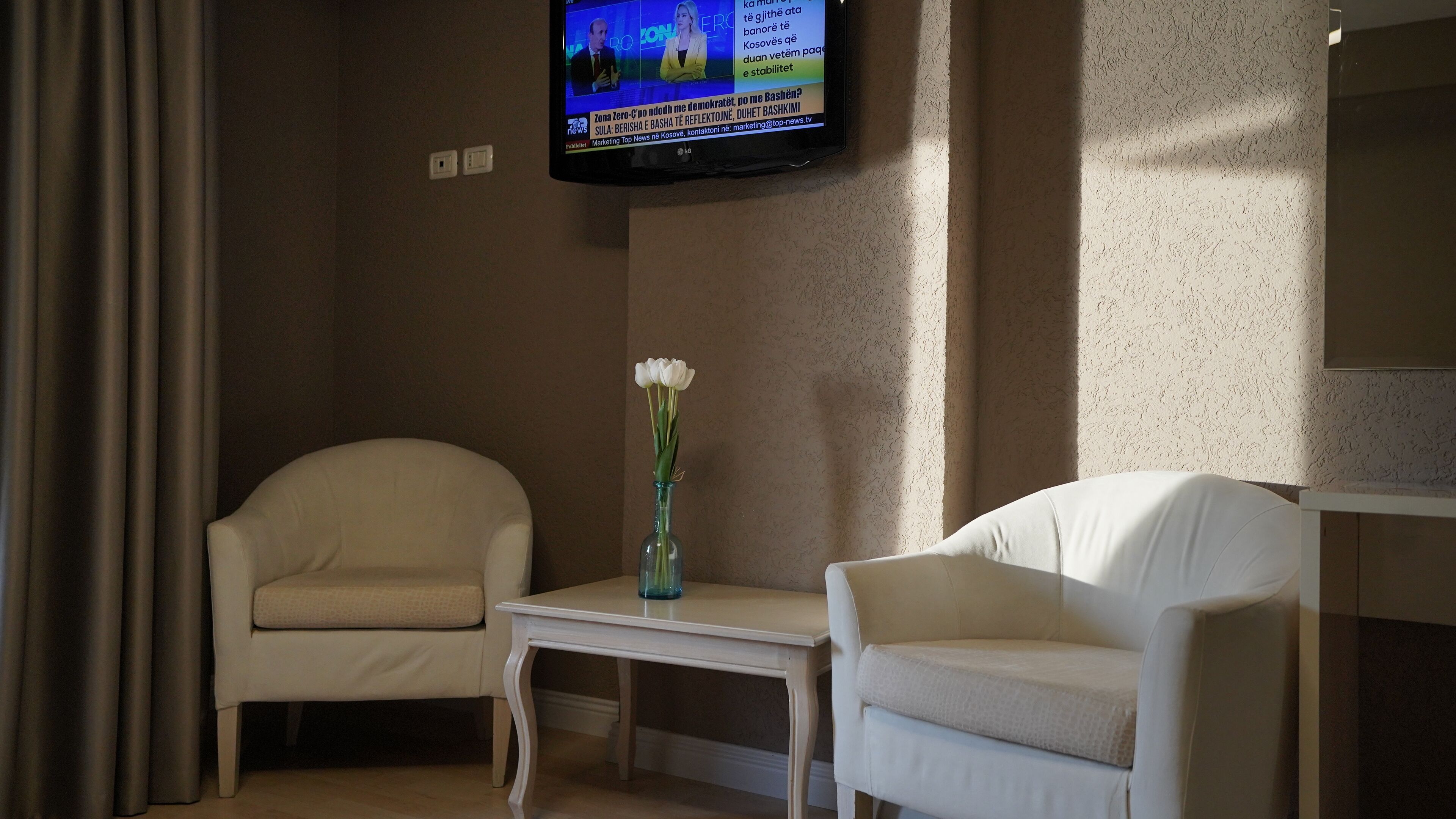 Una camera d'albergo con due poltrone bianche, un tavolino e un televisore fissato alla parete.