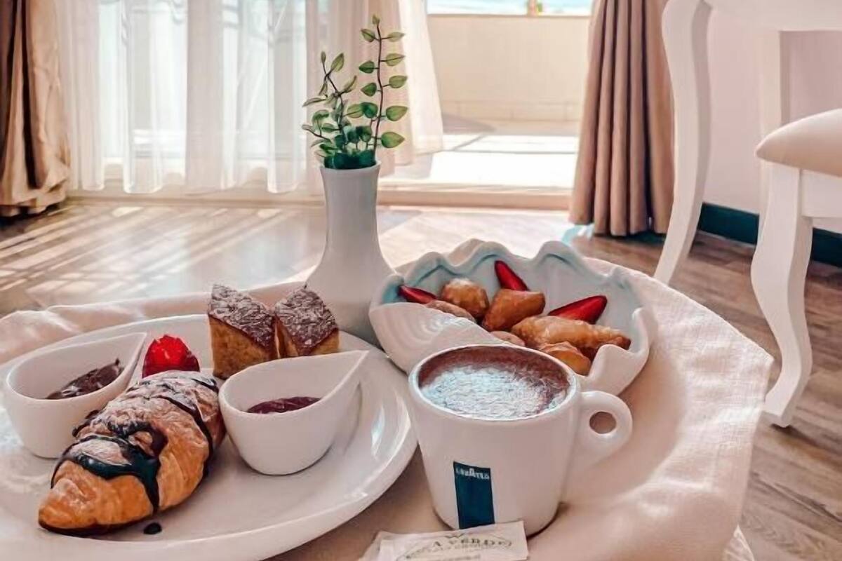 A breakfast tray with pastries, a cup of coffee, and a small plant on a table by a window with curtains.