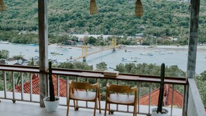 Un balcón con dos sillas de madera y una cactúa en maceta, con vistas a un río con barcos y un puente.