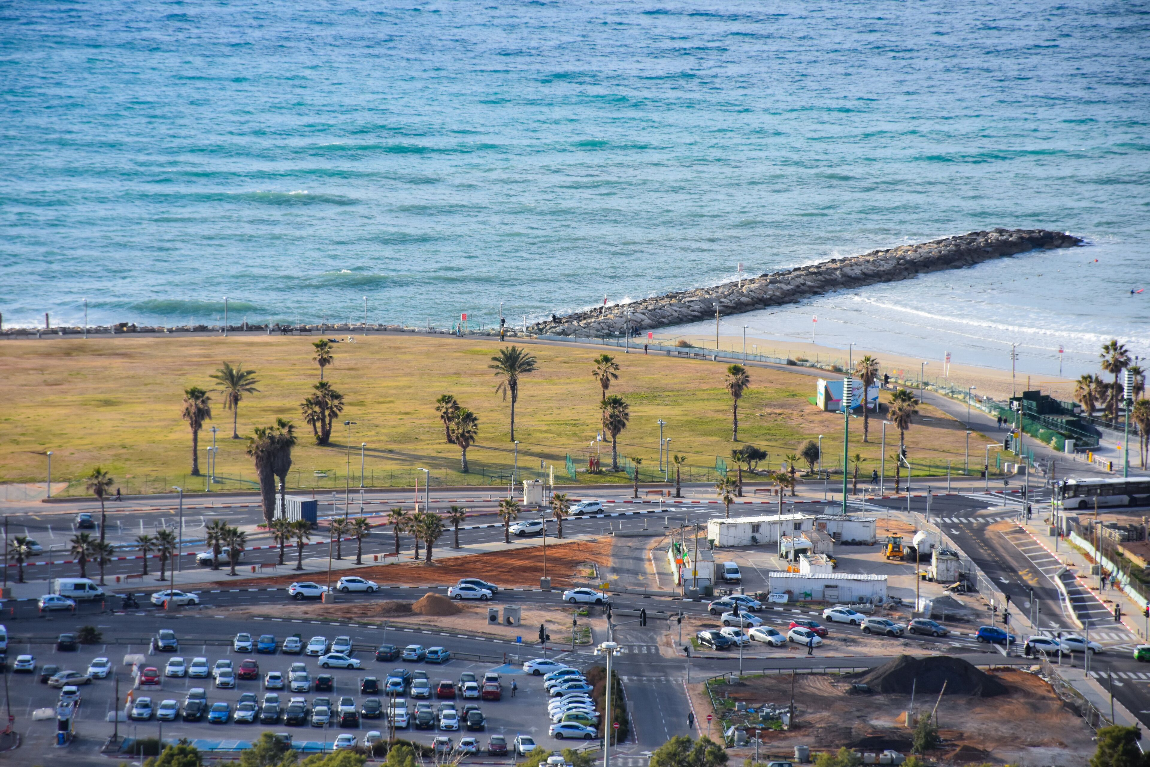 A coastal road with a parking lot, palm trees, and a rocky breakwater.