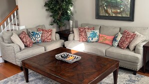 Ein Wohnzimmer mit einem dunkelholznen Couchtisch, einem beige farbenen Sofa mit gemusterten Kissen, einer großen Topfpflanze und einem gerahmten Kunstwerk an der Wand.