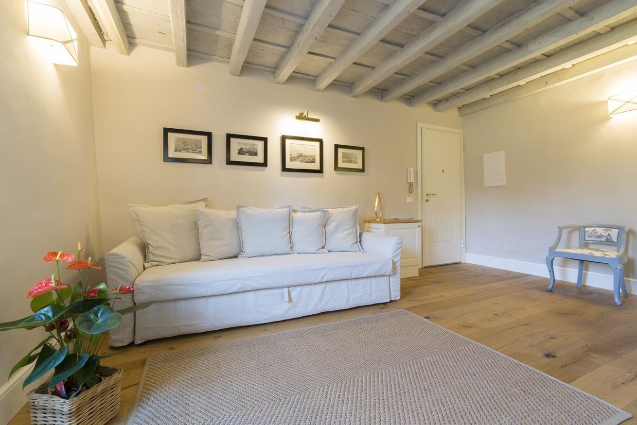 Une pièce avec un canapé blanc, un plancher en bois, un tapis blanc, un luminaire fixé au mur et un tableau encadré.